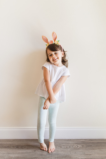 A Happy & Hoppy 4-Year-Old Cuban-American Girl with Brown Curly Hair, Brown Eyes & Olive Skin Wearing a White Shirt, Teal Leggings with Bunnies on Them & Bunny Ears, Eating a Whole Carrot, Barefoot While at Home in the Spring for Easter 2023