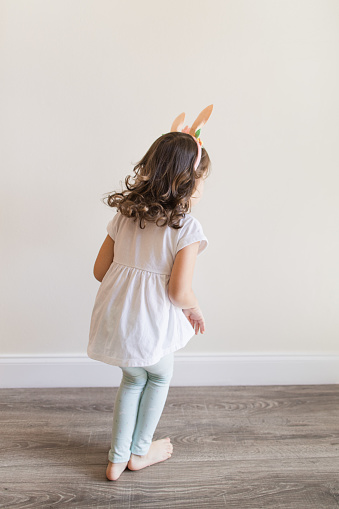 A Happy & Hoppy 4-Year-Old Cuban-American Girl with Brown Curly Hair, Brown Eyes & Olive Skin Wearing a White Shirt, Teal Leggings with Bunnies on Them & Bunny Ears, Eating a Whole Carrot, Barefoot While at Home in the Spring for Easter 2023