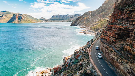 Two cars winding along a scenic road in Kaapstad with majestic mountains and crystal-clear blue water in the backdrop. The perfect view to get lost in!
