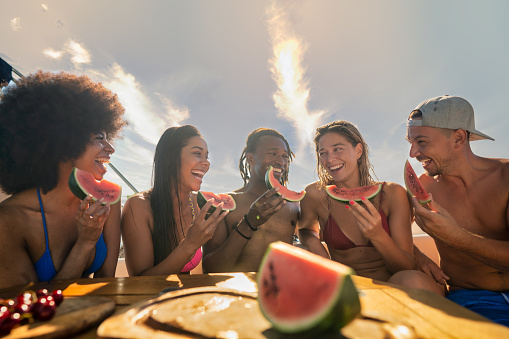 Group of smiling friends on boat at sunset, enjoying slices of watermelon and each other's company. Happiness and relaxation concept