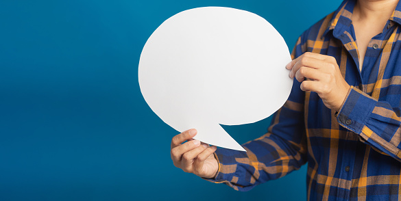 A speech bubble concept. Midsection of a young man holding a blank paper while standing with blue background in the studio. Space for text. Chat and message for social media conceptual ideas