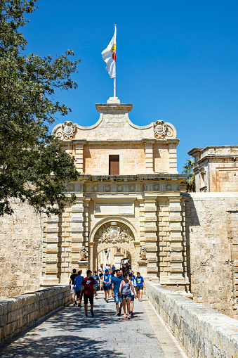 City of Mdina, former capital of Malta, with entrance gate and tourists crossing stone bridge on a hot summer day. Photo taken August 8th, 2017, Mdina, Malta.