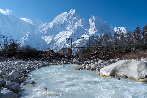 A flimsy bridge over a small mountain river, consisting of thin metal tubes. Old pedestrian bridge over the river against the background of snow-capped mountain peaks