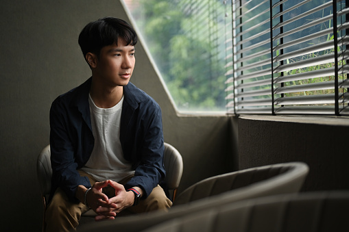 A handsome Asian man in a casual shirt is contemplating and looking out the window.