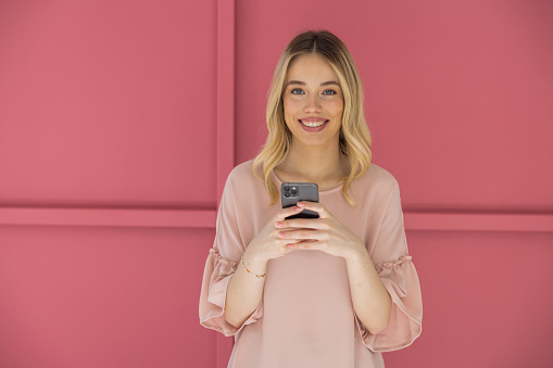 Young blonde woman on pink background using smart phone.