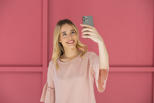 Young blonde woman on pink background using smart phone.