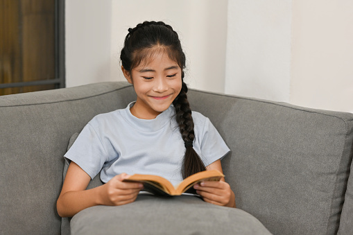 Happy cute little girl reading book on sofa at home, Education and Learning concept.