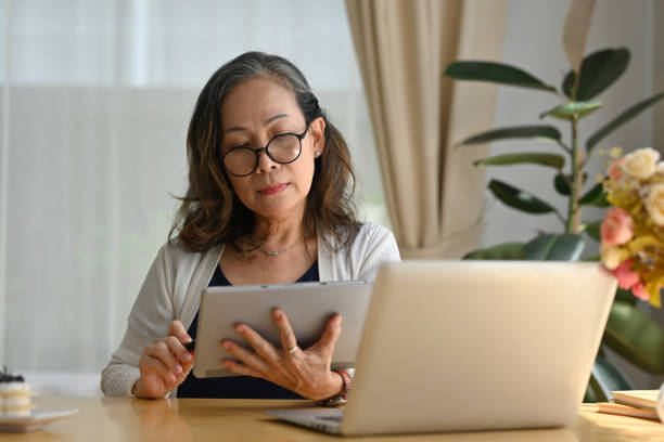 istock Stylish mature old woman remote working from home, using digital tablet and laptop for working and watching webinar. 1486227014