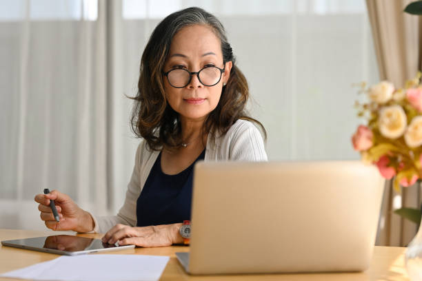 istock Smiling 60s middle aged business lady remote working from home, using laptop watching webinar and taking notes on digital tablet. 1486227015