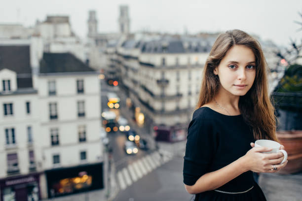 Young pretty dreamy woman looking at camera and standing with cup on balcony in Paris, France.