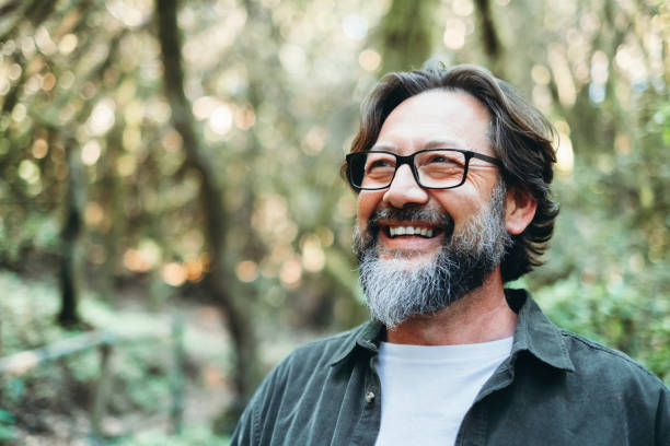 Portrait of young mature man alone smiling outside with nature trees in background. Environment. Happy people enjoying outdoor leisure activity. Nature feeling. Bearded adult male wearing eyeglasses stock photo