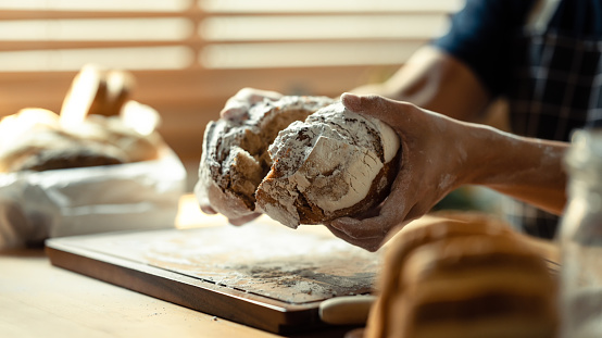 Close up view of man hands breaking fresh bread, sitting at table with flour and kitchen utensils.