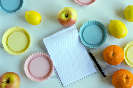 An open paper notebook and pen on a white kitchen table among fresh fruits and a variety of nuts. The concept is the compilation of a menu, a list of products, and the design of recipes.