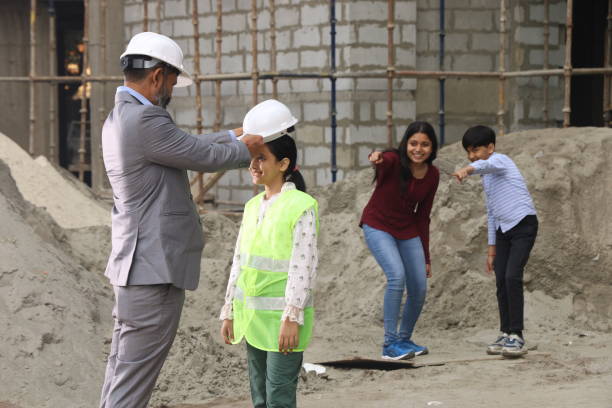 istock Children visit on a construction site. Hardworking Indian Site engineer entertaining kids and showcasing them the construction work. On the site location wearing the uniform and helmet in white. Quality control and inspection. 1486241601