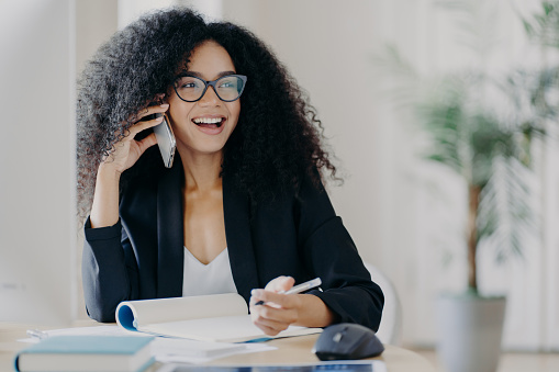 Photo of glad smiling dark skinned woman calls business partner, being in good mood, makes notes, sits at workplace, wears optical spectacles, formal suit, poses in cabinet. Work, business, technology