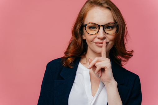 Isolated shot of attractive secret woman keeps fore finger over lips, demonstrates shush gesture, tells secret information, has healthy skin, manicure, makeup, wears formalwear, poses over pink wall