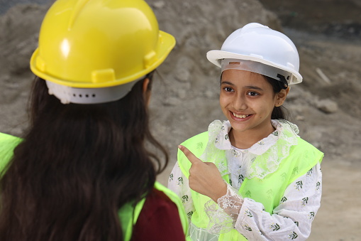 Young Indian School kids working as a role model engineer on a builder construction site working on a blueprint of the property. They are  wearing construction engineer helmet.