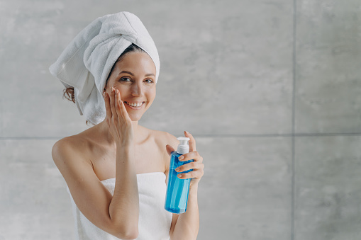 Face toner or micellar water advertising concept. Happy young woman touches pure skin. Gorgeous caucasian woman wrapped in towel after bathing applying toner in flacon. Face washing foam in bottle.