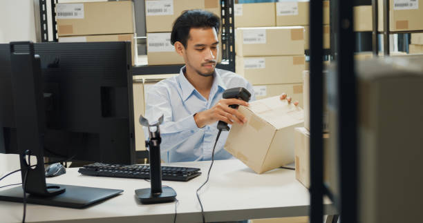 istock Happy Asian man wear formal uniform shirt sit in front of desk use computer and barcode machine scanner record customer online data in cardboard box delivery at warehouse. Startup business. 1486253461