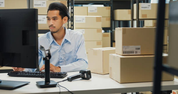 istock Happy Asian man wear formal uniform shirt sit in front of desk use computer and barcode machine scanner record customer online data in cardboard box delivery at warehouse. Startup business. 1486253468