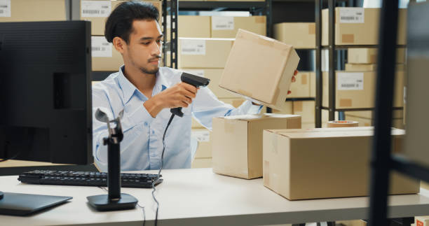 istock Happy Asian man wear formal uniform shirt sit in front of desk use computer and barcode machine scanner record customer online data in cardboard box delivery at warehouse. Startup business. 1486253487