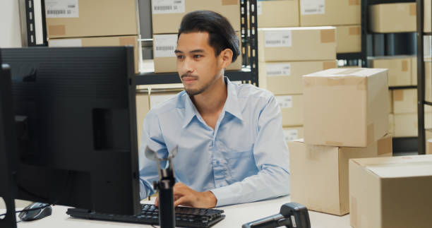 istock Happy Asian man wear formal uniform shirt sit in front of desk use computer and barcode machine scanner record customer online data in cardboard box delivery at warehouse. Startup business. 1486253496