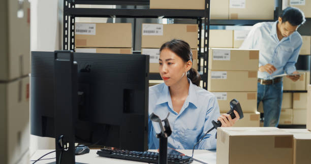istock Happy Asian girl and man partner wear formal shirt sit front desk computer use barcode machine scan record customer online data in cardboard box delivery at warehouse. Startup small business. 1486253521