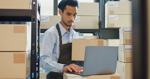 Asian business partner man wear formal shirt hold cardboard box use laptop computer check inventory online stock data program for delivery customer at warehouse. Startup small business. stock photo