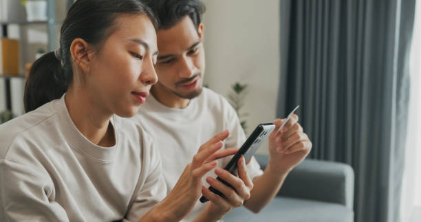 istock Happy young Asian couple sit on couch spend time together have fun use smartphone devices shopping use credit card online payment at home. Young husband and lifestyle domestic activity. 1486256251