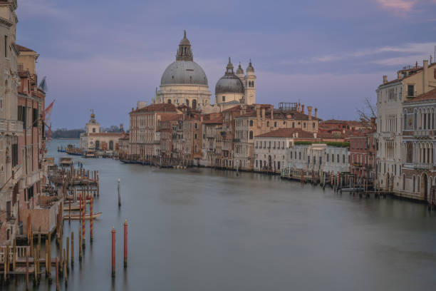 People sitting on the steps at the entrance of Santa Maria della Salute church (Saint Mary of Health) by the Grand Canal at Venice, Italy. The Grand Canal is an ancient waterway that goes from the Saint Mark Basin to a lagoon near the Santa Lucia rail station.\nThe city of Venice is an Unesco World Heritage site and it is visited daily by thousands of tourists from all over the world. Some gondolas are floating next to the sidewalk.