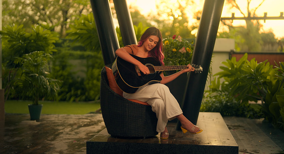 Beautiful Indian woman sitting on chair playing acoustic guitar and singing alone outside home. Happy Cheerful female enjoy evening entertainment sing song new sound recreation on classical lesson Learning course, practicing as hobby avocation