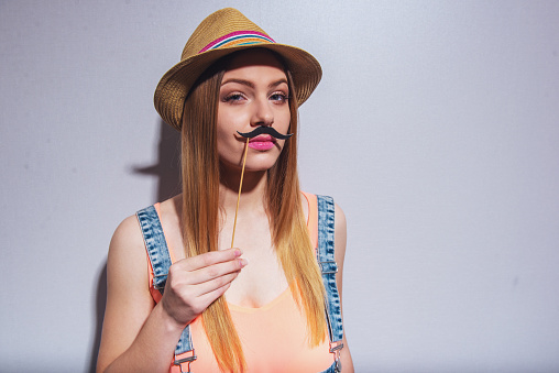 Attractive girl in summer clothes is posing with paper moustache on wood stick and looking at camera, against gray background