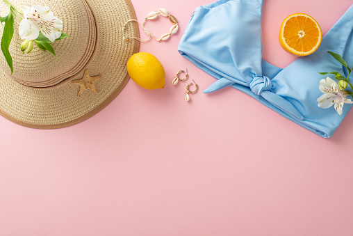 Ready for the beach! Top view flat lay of blue swimsuit with sunhat, orange fruit lemon, earrings, bracelet, and flowers on pastel pink background with empty space for text or advert