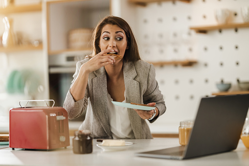 Shot of a multi-tasking young business woman using laptop in her kitchen and having a breakfast while getting ready to go to work.