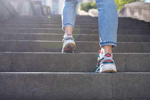 Close-up of female tourist in casual clothes going upstairs outdoors. Woman in jeans and sneakers going up steep stairs