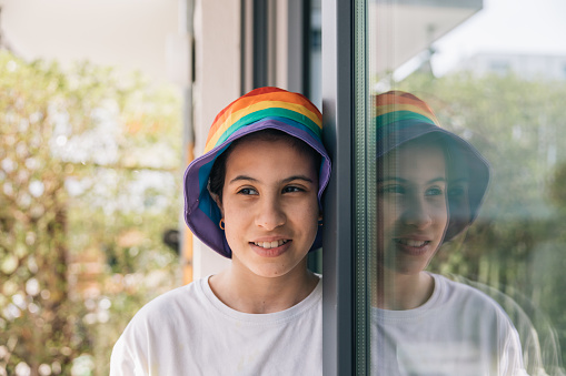 Teenager with rainbow hat smiling reflected in window, LGBTQ+ Pride Month concept