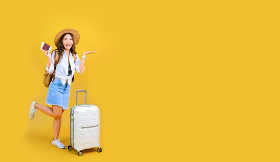 young Asian pretty woman traveling abroad smiling and looking at camera isolated on yellow studio background, cheerful passenger pointing while standing with valise suitcase during summer vacation