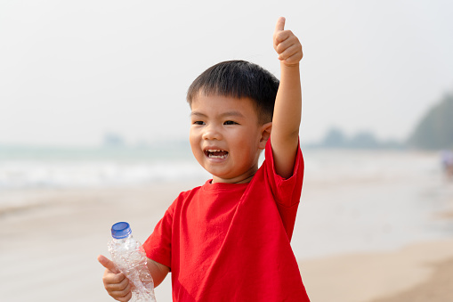 happy little boy collects trash drink water bottle for recycling and raises arm thumb-up, volunteer kid cleaning sea sand beach for protecting environment from ocean problems sustainable future