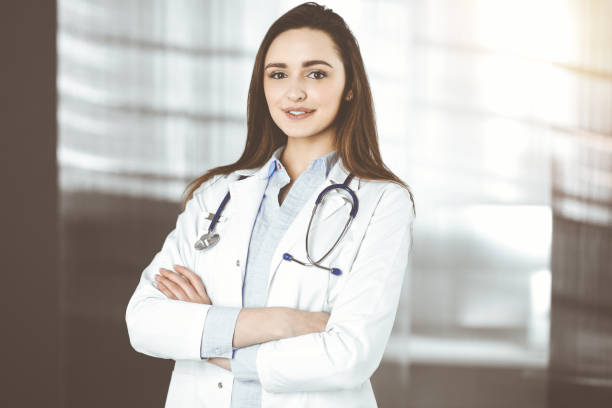 une jeune femme-médecin souriante se tient debout dans une clinique ensoleillée à l’intérieur - clothing professional occupation medical occupation doctor photos et images de collection