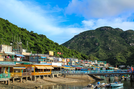 The view of the beach of Cheung Chau island of Hong Kong with mountains in the background under blue sky. Colorful village houses in the coastline with boats besides the coast. Travel and nature scene.