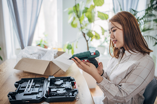 Young Asian woman opening a cardboard package, while sitting at the table at home, she ordered a glue gun work tool.