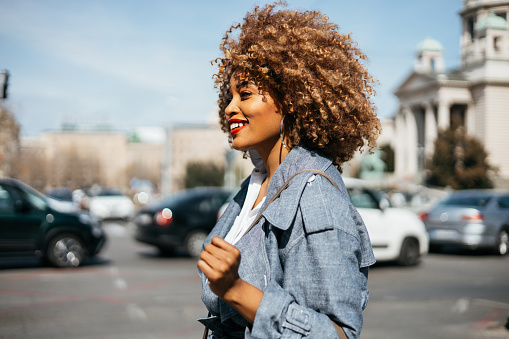 Beautiful black fashionable woman walks down the street. She is happy and smiled. Bright sunny day. Side view.