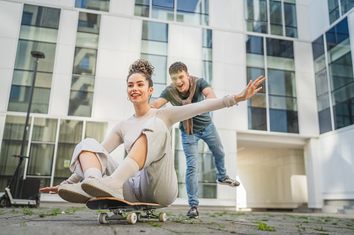 Young man and woman teenager couple caucasian brother and sister have fun on skateboard pushing to ride by the modern building in day happy smile real people family love concept copy space