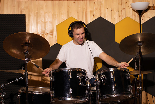 White Caucasian man emotionally plays on drums with ear pads in underground studio. Soundproofed wooden wall on background. Darken photo. Dynamic musician playing with sticks. Horizontal plane.