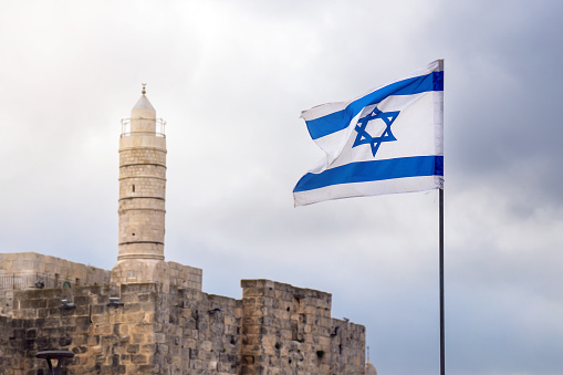 Israeli flag flutters in the wind with a muslim minaret of a mosque on the background in Jerusalem in cloudy sky