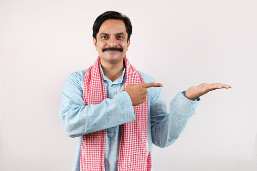 Portrait of an happy Indian farmer in rural India concept. Standing in white background, the cheerful farmer is pointing finger towards the other hand. Excited farmer is very joyous and exuberant