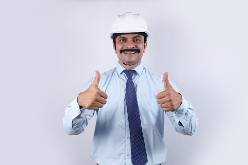 Portrait of a happy Indian engineer wearing an engineer helmet white in color. He is confident and showing a thumbs up.