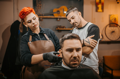 Young beautiful female barber with short red hair learning to cut hair in. She is in education training in a barber shop. A young handsome male barber observes her work.