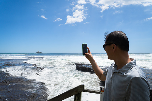 Tourist taking smartphone photos of big waves crashing onto rocks at Muriwai Beach. Auckland.