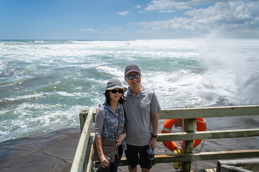 Tourists posing for photos at Muriwai Beach. Big waves crashing on to rocks behind them. Auckland.